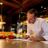 Chef looks at iPad while jotting down on a notepad. Photo.