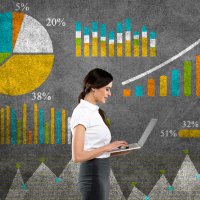 Photo of a woman standing in front of some graphs and tables, holding a laptop.